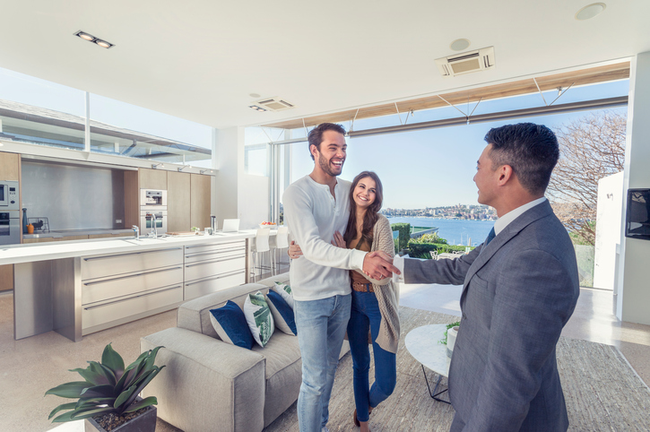 Real estate agent with couple in luxury home. They are shaking hands. There is a water view, kitchen and living room in the background. Couple are casually dressed. They are laughing. Agent is dressed in a suit and smiling. Wide angle.